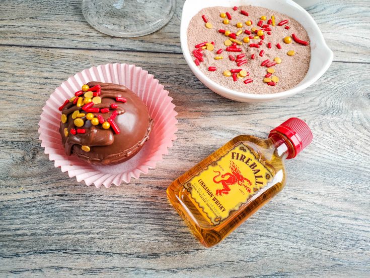 hot chocolate bomb sitting in a cupcake liner, topped with red and gold sprinkles. A small bottle of fireball whiskey, and hot cocoa in a white bowl all sitting on a wood table