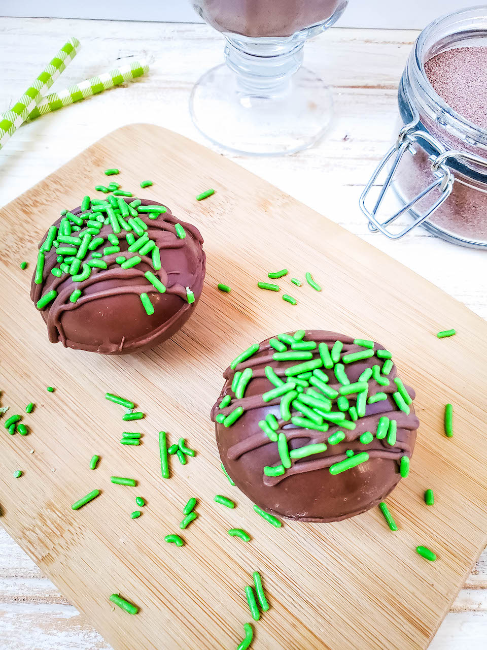 two cocoa bombs sitting on a wooden board covered with green sprinkles. Green stripped straws and a jar of cocoa mix in the background