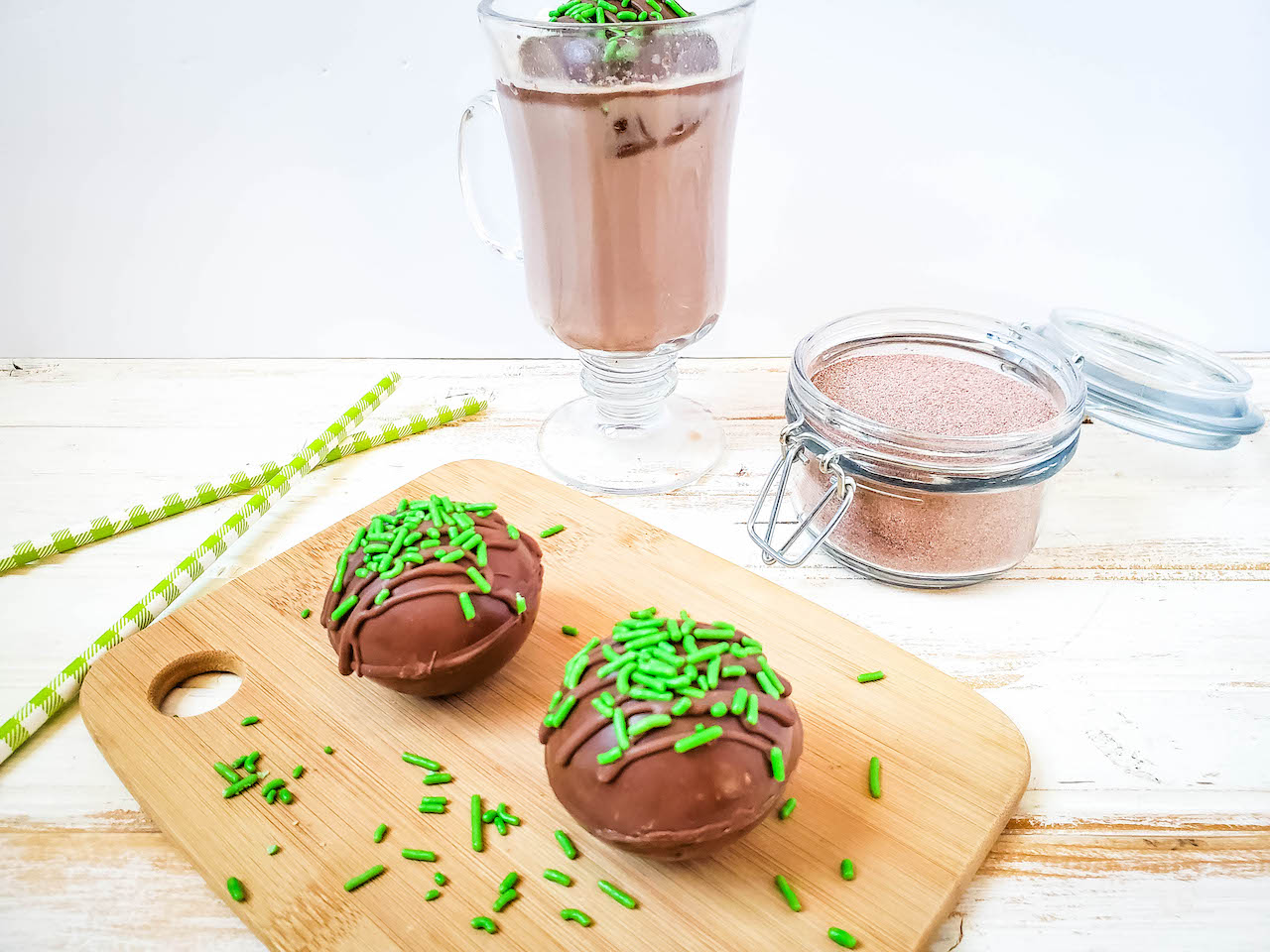 two hot cocoa bombs sitting on a wood cutting board with green sprinkles. A cup of hot cocoa and a glass jar of hot cocoa mix in the background, with two green striped straws