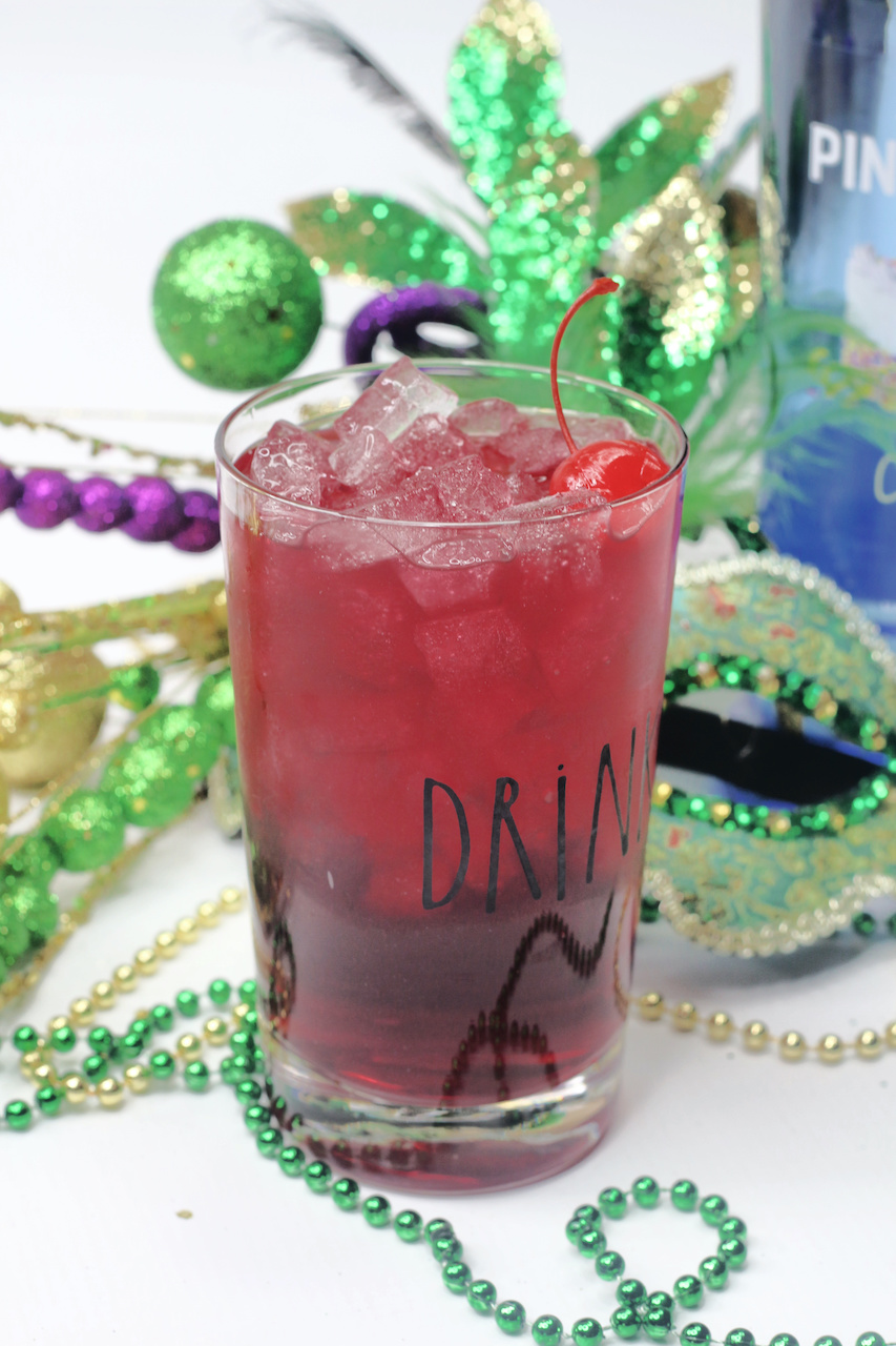 a clear glass filled with cocktail sitting in front of Mardi Gras inspired decorations