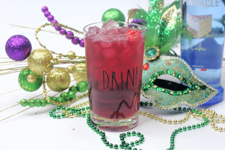 a clear glass filled with King Cake Cocktail sitting in front of beads, a mask and other Mardi Gras decorations