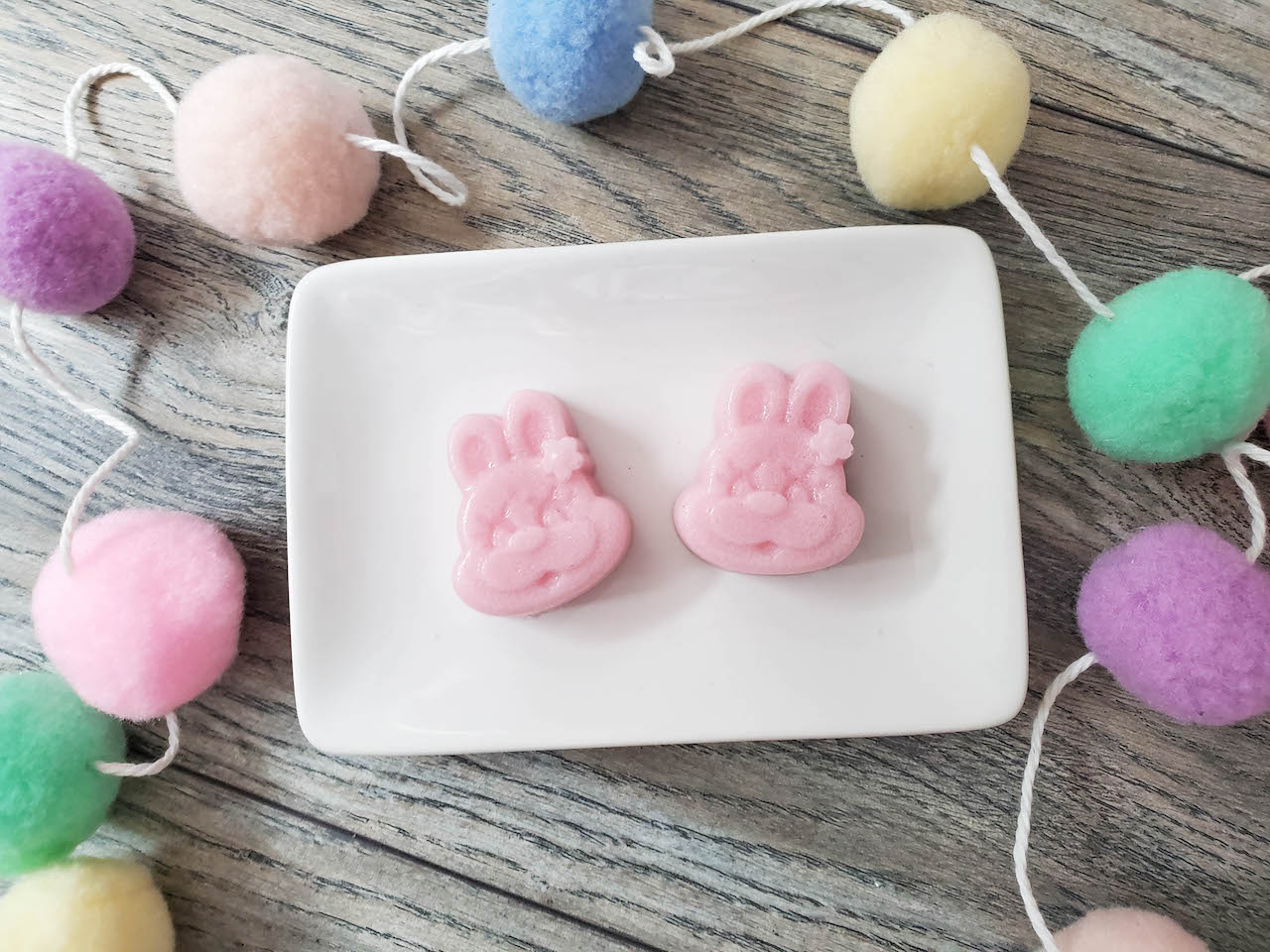 two bunny face diy sugar scrub bars sitting on a white rectangular plate surrounded by easter Pom Pom garland strand