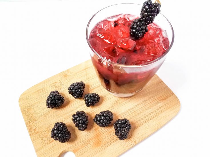 a glass of blackberry margarita garnished with fresh blackberries sitting on a wooden cutting board with additional whole fresh blackberries