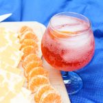 an easy summer cocktail in a glass sitting on a blue cloth next to a cutting board and knife with orange slices, cheese slices, and strawberries