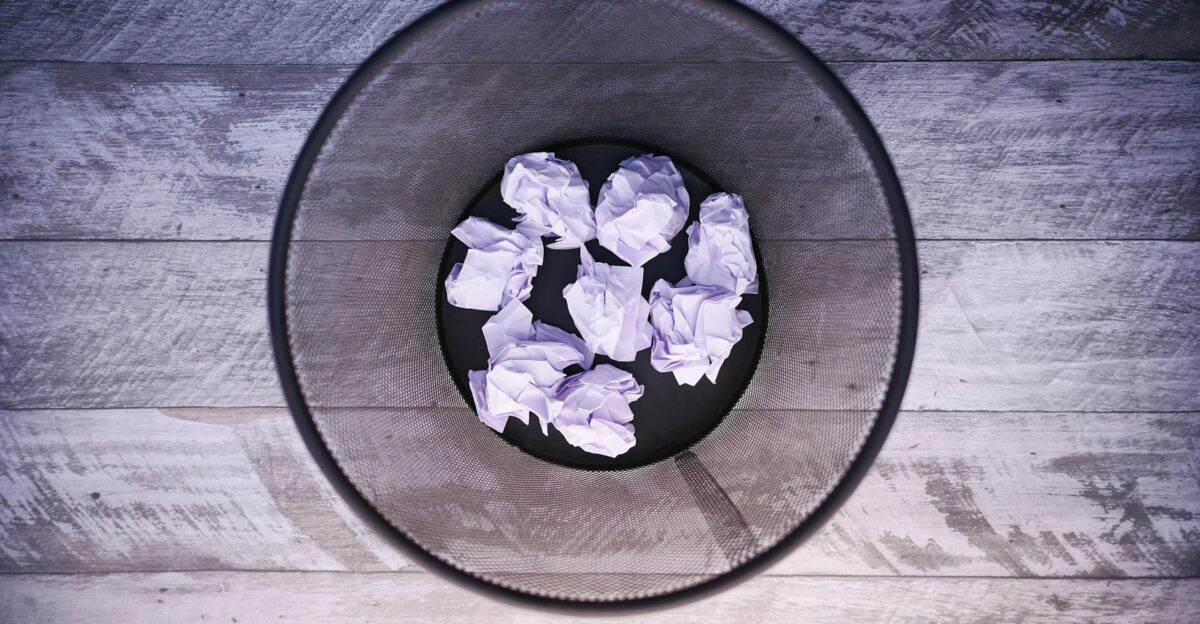 A top-down view of crumpled papers inside a black trash can on wooden flooring