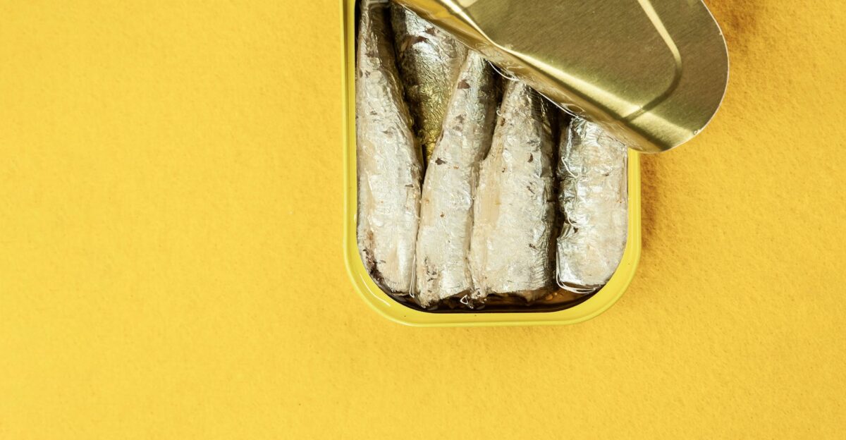 Top view of canned sardines with an open lid on a yellow surface showcasing food preservation