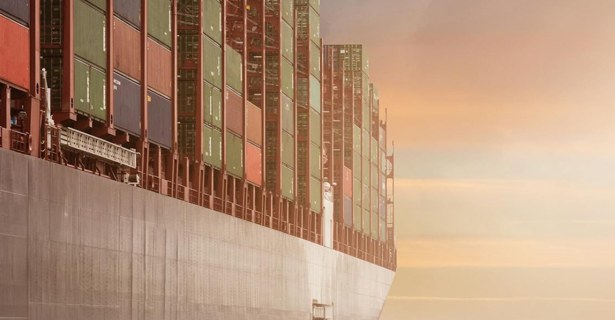 A colossal cargo ship loaded with containers navigates through calm waters against a vibrant sunset sky.