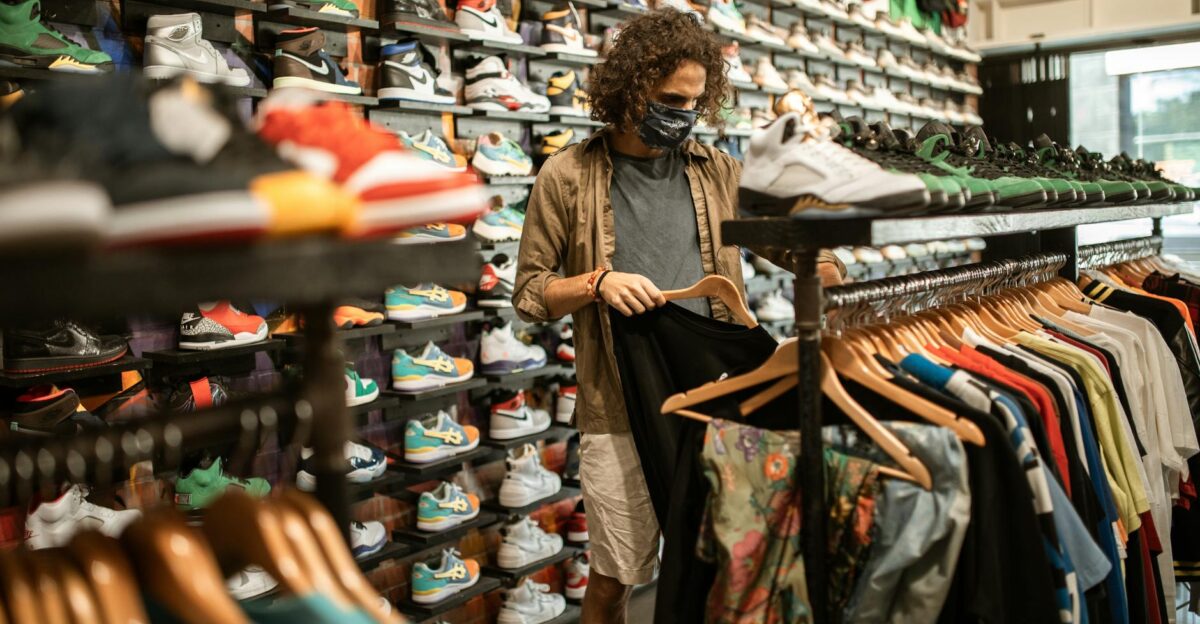 Young man in a sneaker and clothing store wearing a mask browsing through shirts amid pandemic