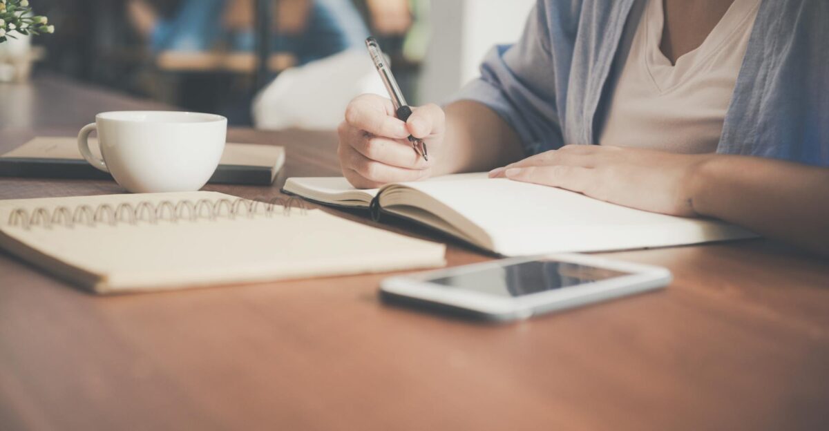 A woman writes in a notebook at a caf table with a coffee and smartphone nearby