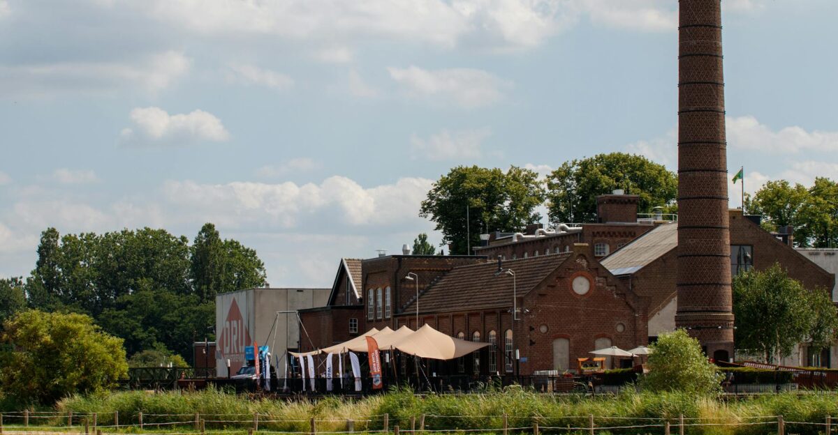 View of an old brick factory with a tall chimney surrounded by greenery and a partly cloudy sky