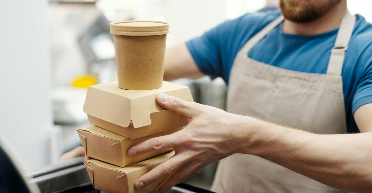 Man wearing apron stacks eco-friendly food containers for takeaway delivery in a kitchen setting