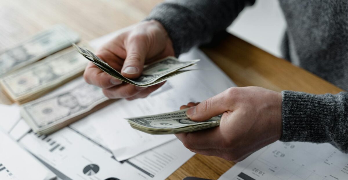 Person counting dollar bills on a desk with financial documents and a calculator in the background
