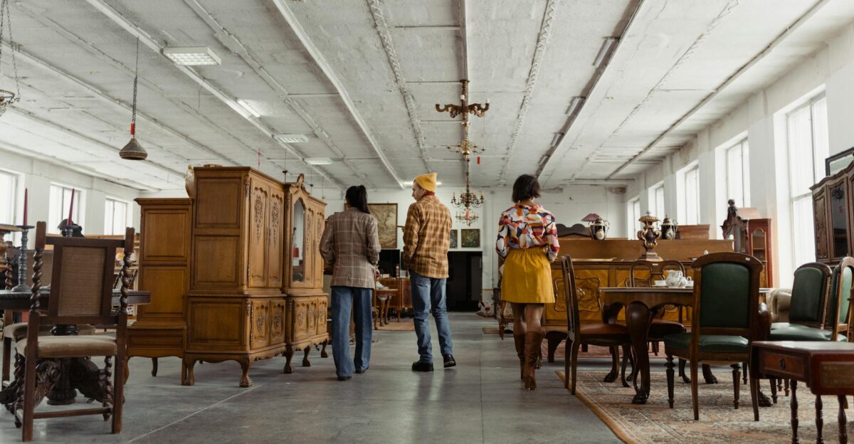 Three people browsing vintage wooden furniture in a spacious store interior with high ceilings