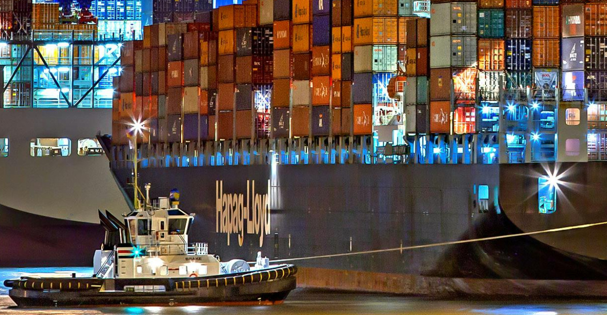 A brightly lit cargo ship at Hamburg harbor with stacked containers and a tugboat.