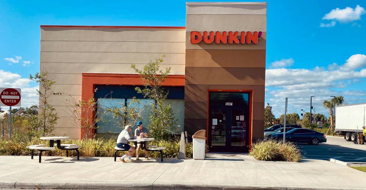 Exterior of a Dunkin store with people sitting outdoors under clear blue sky